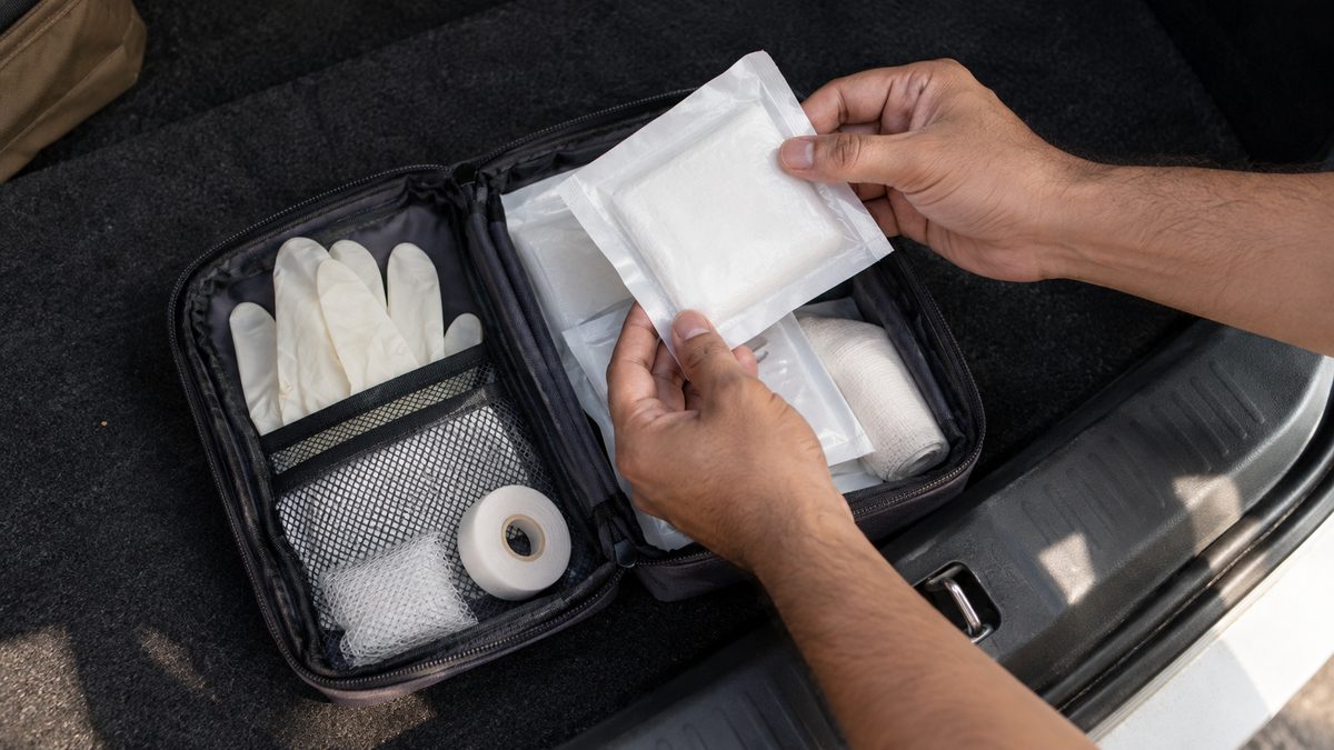 Hands checking sealed dressing packets in an open first aid pouch on a car’s boot floor.