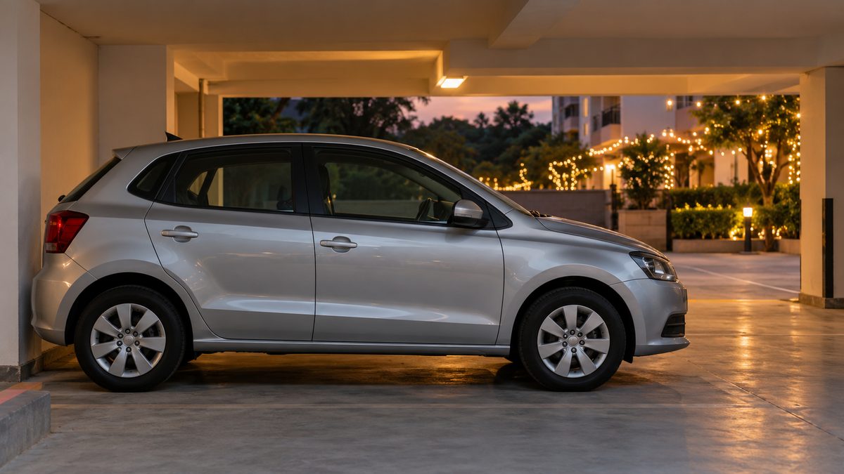 A silver car parked under a residential building in India with festive lights outside at dusk.