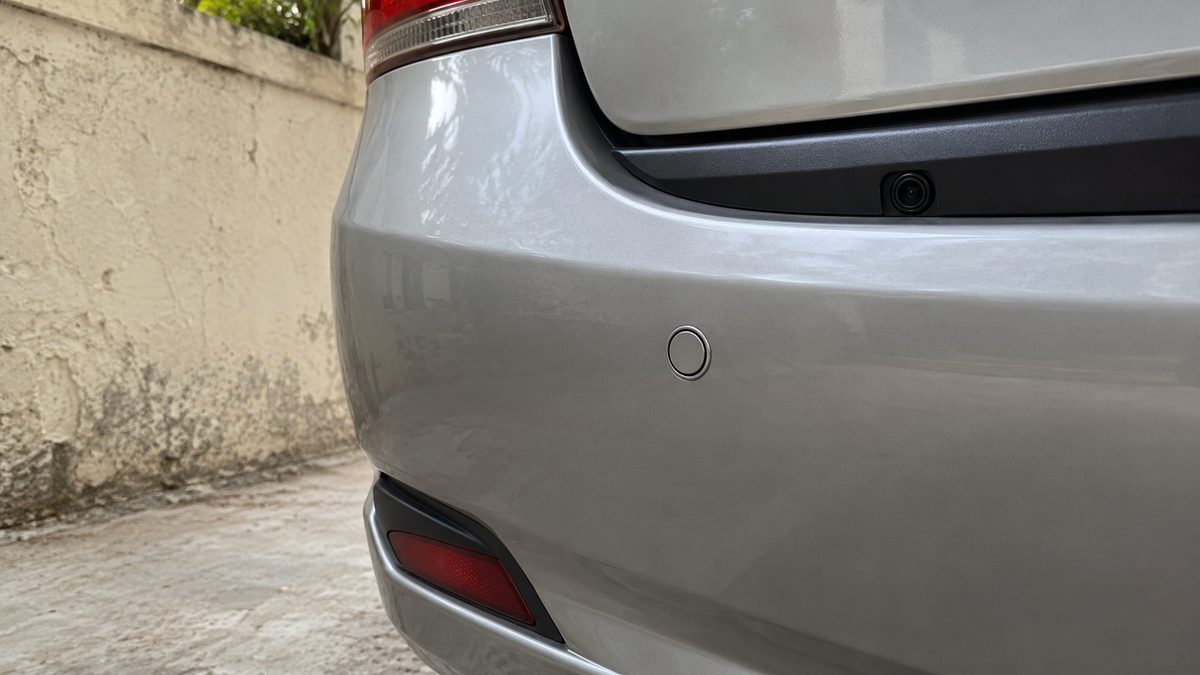 Small reversing camera and circular parking sensor fitted to a silver rear bumper beside a plaster wall.